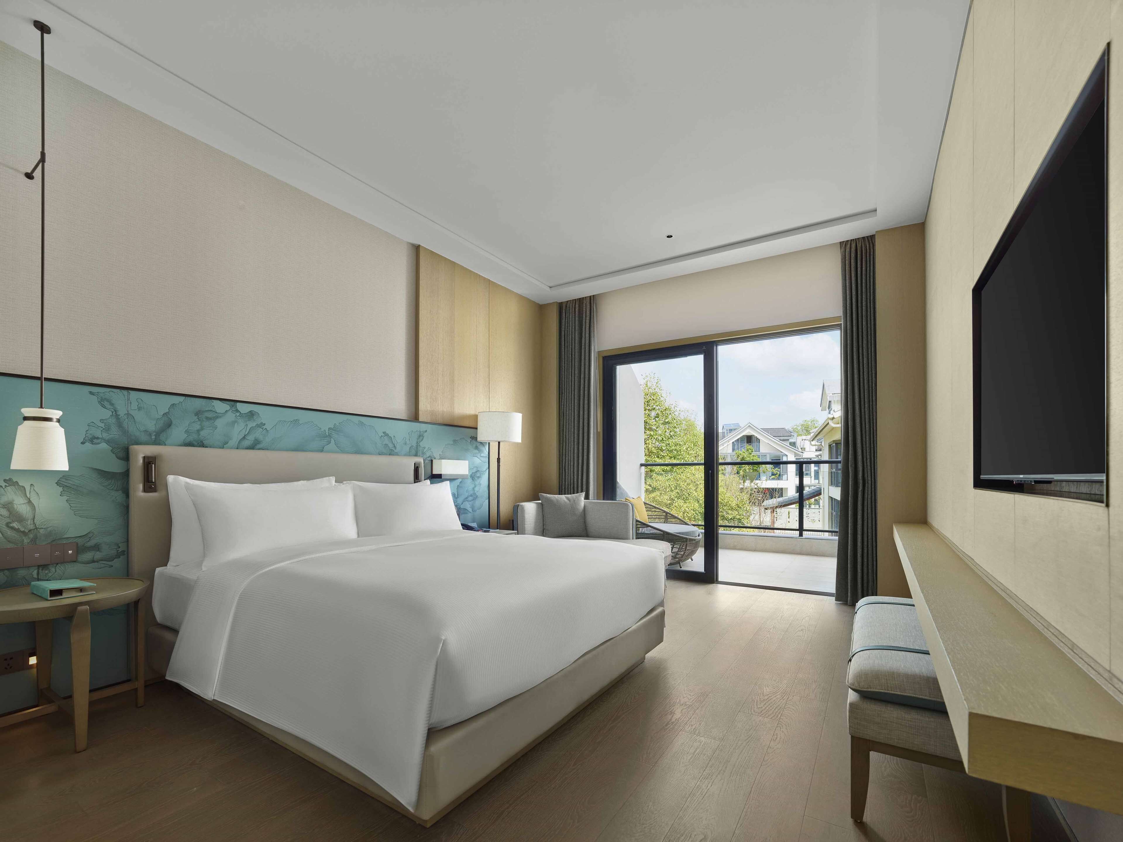A hotel room with a large bed, a desk, a TV, and a balcony with a view.