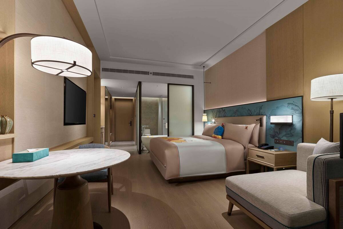 Een moderne hotelkamer met een groot bed, nachtkastjes, een bureau met stoel, een ronde tafel en een groot raam.