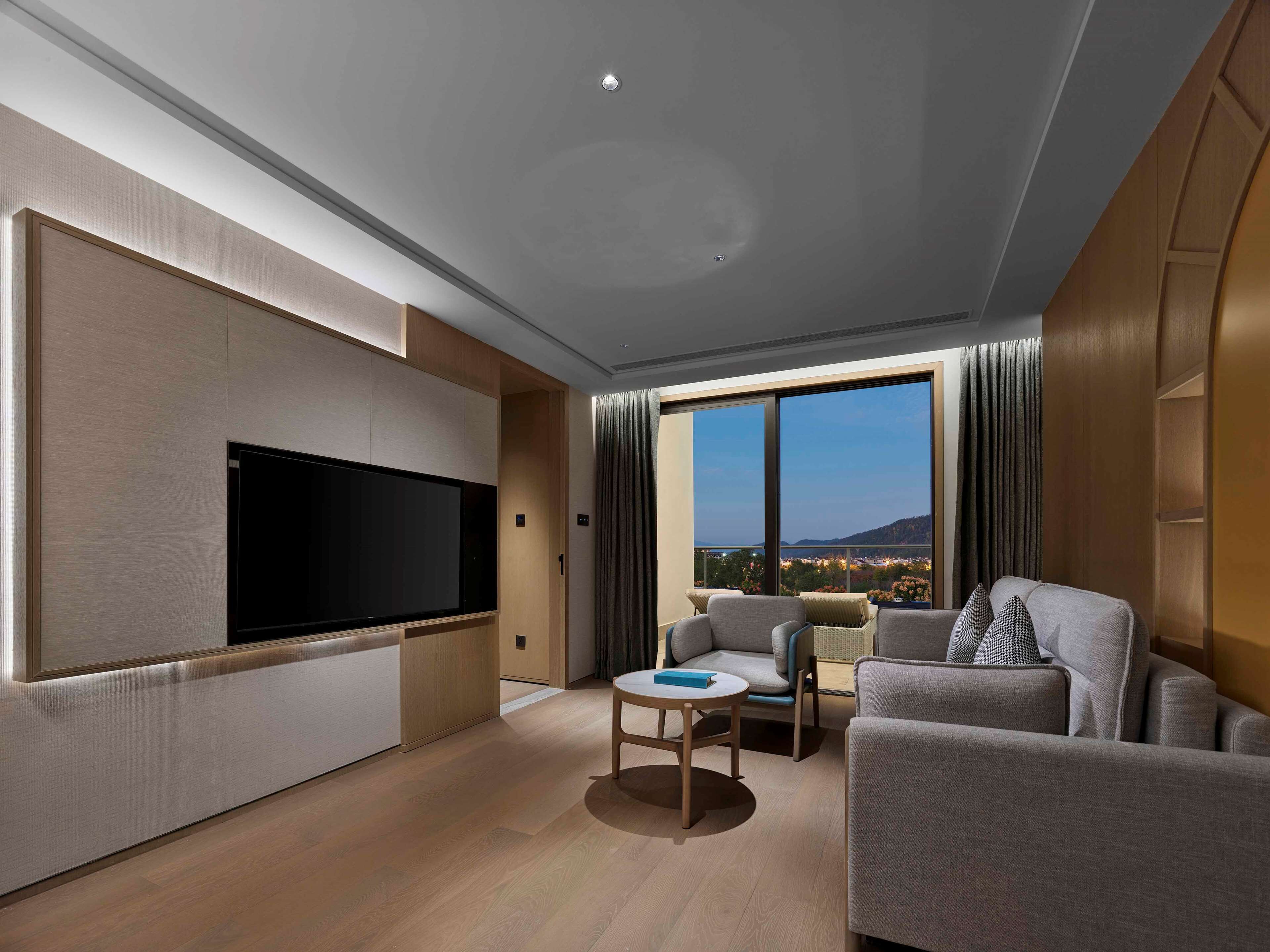 Una camera d'albergo moderna con una grande TV a schermo piatto, un divano, un tavolino e un'ampia finestra con vista sulla città.