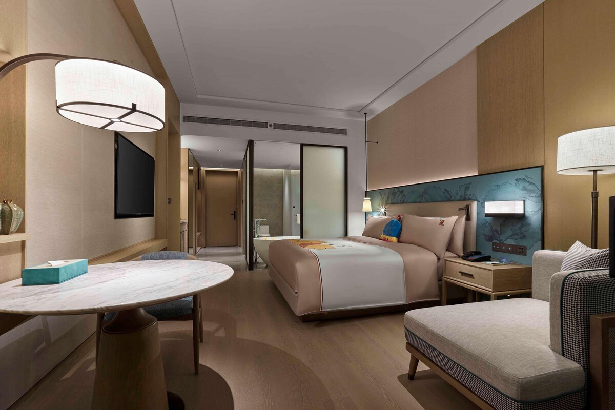 A modern hotel room with a large bed, a desk, and a chair. There is a TV mounted on the wall and a window with a view of the city.