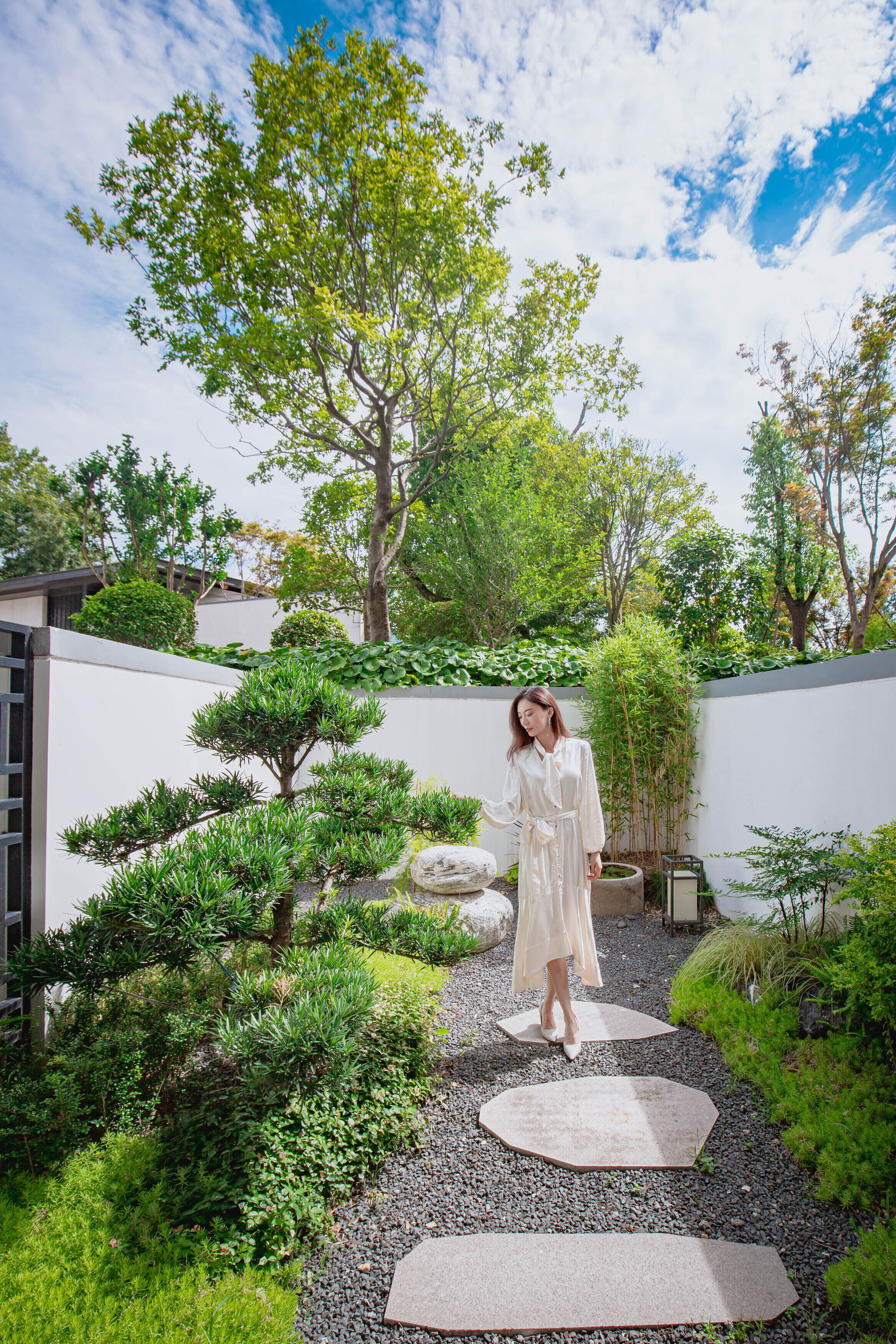 A woman walking on a stone path surrounded by lush greenery and trees in a garden setting.