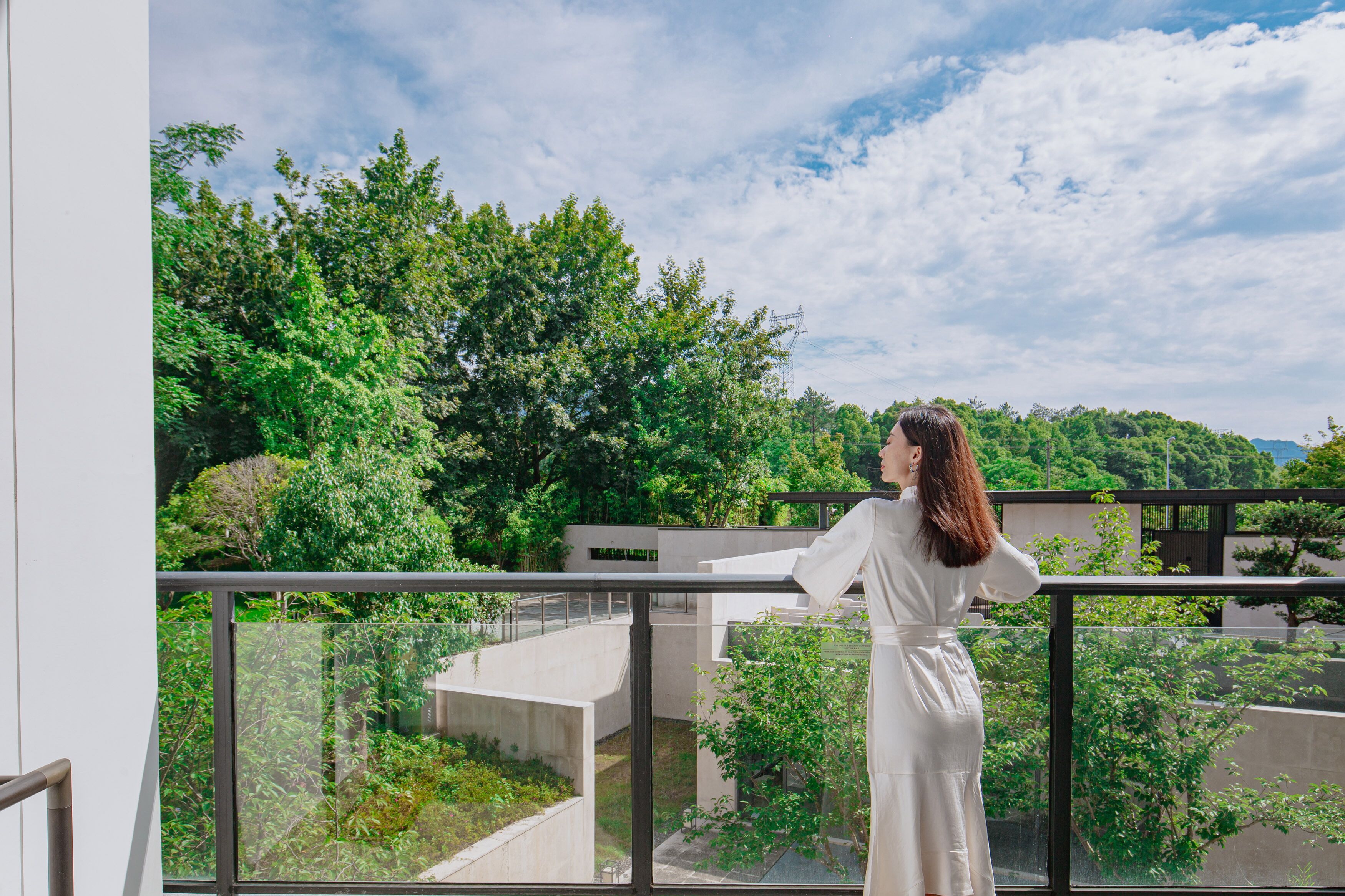 Eine Person steht auf einem Balkon mit Blick auf Grünflächen und Gebäude.