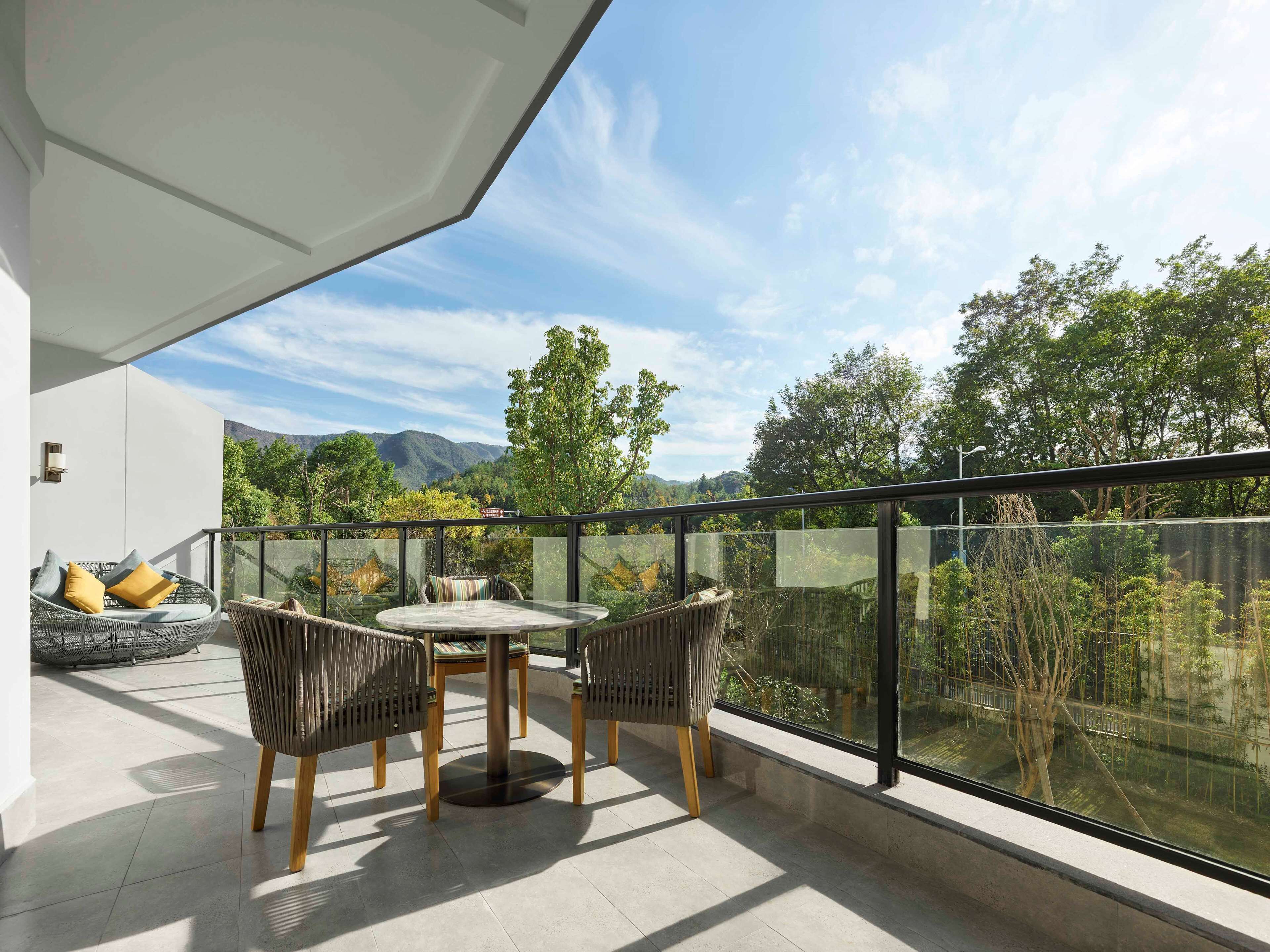 Un balcone con ringhiera in vetro, mobili in vimini e vista su alberi e montagne.