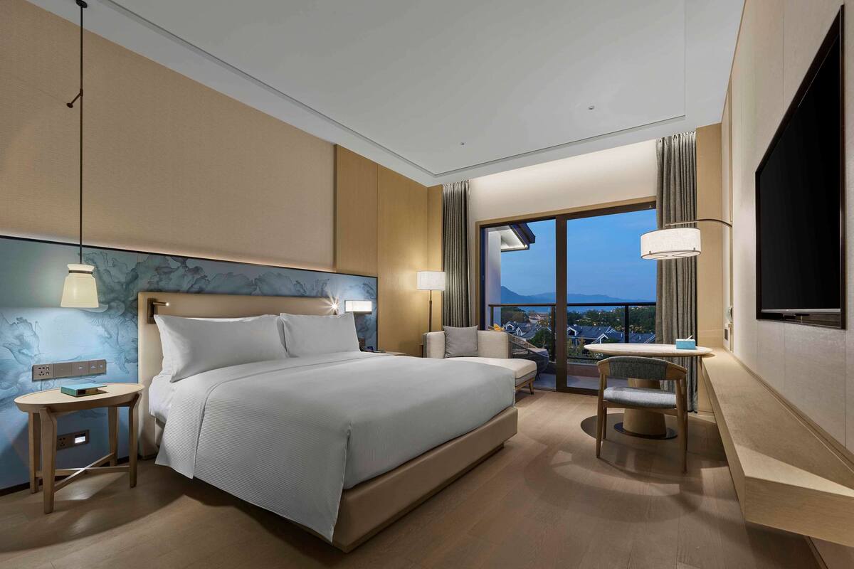 Een hotelkamer met een groot bed, nachtkastjes, een bureau met stoel, een televisie en uitzicht op de stad door grote ramen.
