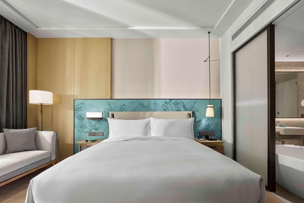 A hotel room with a large bed, a sofa, a nightstand with a lamp, and a bathroom with a mirror and sink.