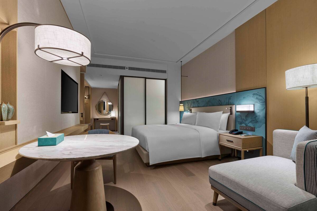 Een moderne hotelkamer met een bed, nachtkastje, lamp en een ronde tafel met een zakdoosje.