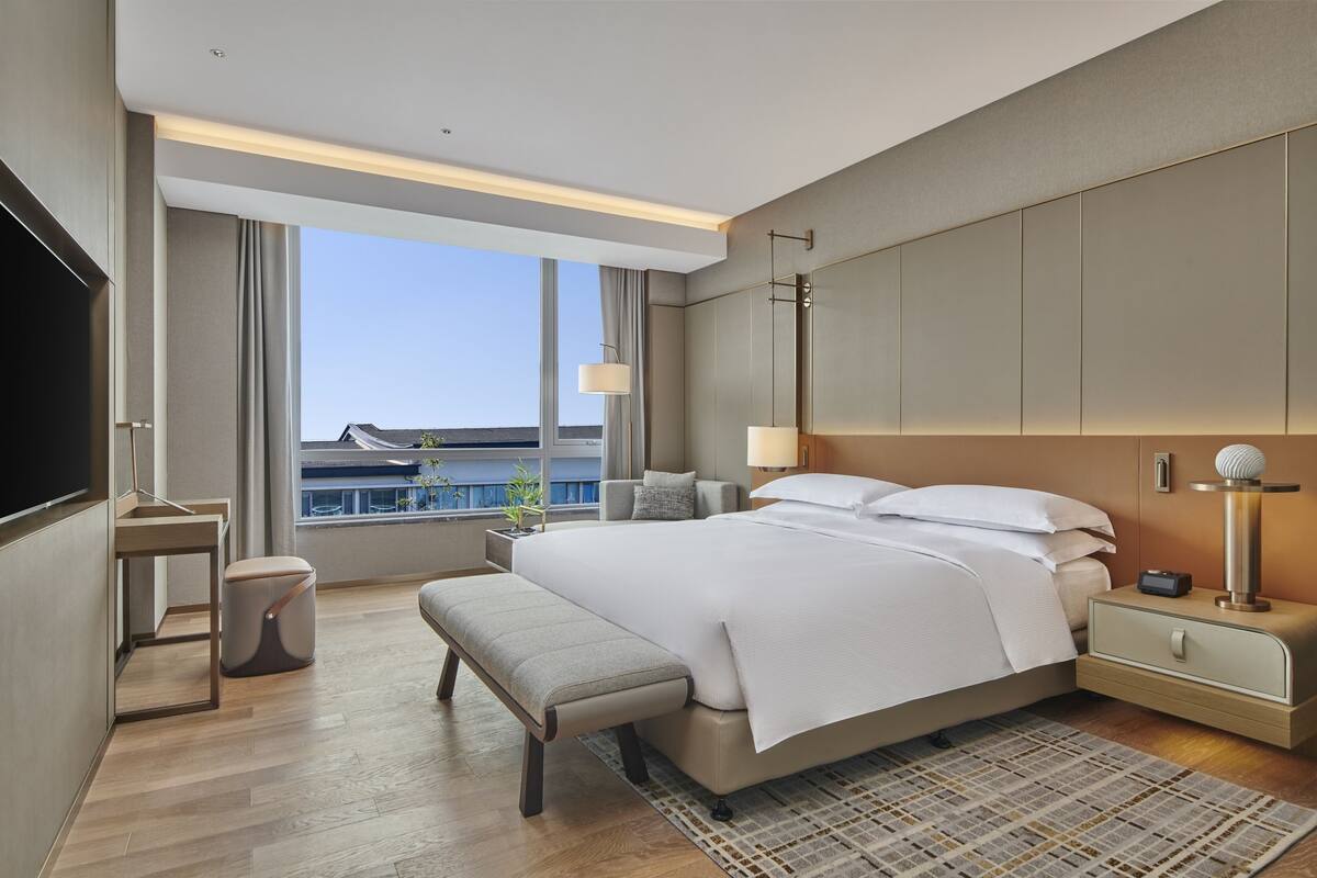 Une chambre d’hôtel moderne dotée d’un grand lit, de tables de chevet, d’un banc, d’une télévision et offrant une vue sur la ville.