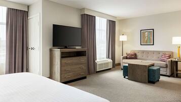 Habitación de hotel con una cama grande, un televisor sobre un soporte de madera, un sofá, una mesa de centro y una ventana con cortinas.