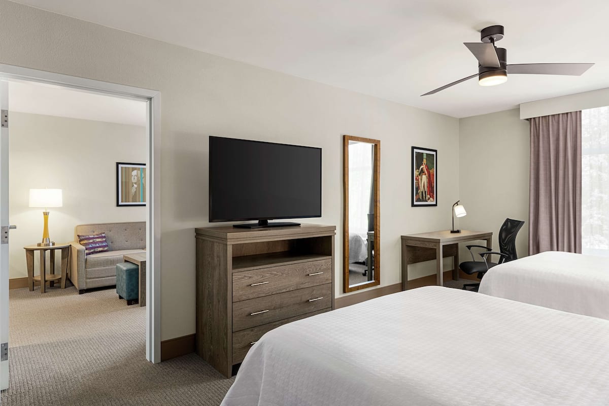 Ein Hotelzimmer mit zwei Betten, einem Fernseher, einem Schreibtisch mit Stuhl, einem Spiegel und einem Deckenventilator.