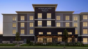 Un hôtel à plusieurs étages au design moderne, avec une enseigne bien visible indiquant « Homewood Suites by Hilton » et une entrée bien éclairée dotée d’un espace extérieur aménagé avec des sièges.