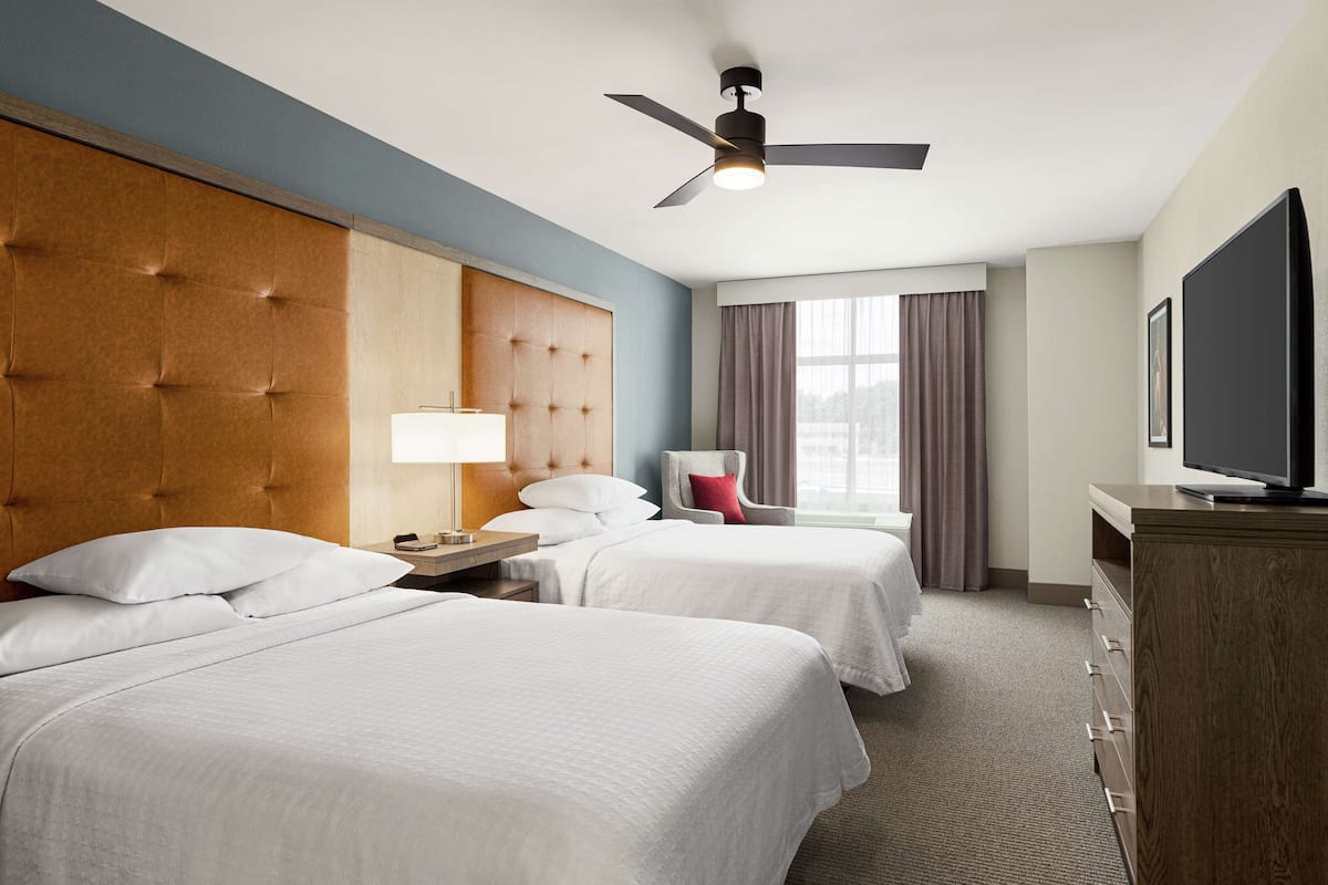 Ein Hotelzimmer mit zwei Betten, einem Deckenventilator, einem Fernseher, einem Schreibtisch mit Lampe und einem Fenster mit Vorhängen.