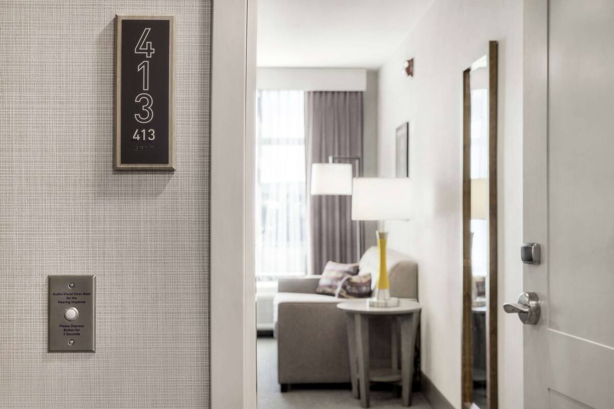 Ein Hotelzimmer mit Hinweisschild, einem audiovisuellen Türsignal, einem kleinen Tisch mit Lampe und Blick auf einen Wohnbereich mit Sofa und Lampe.