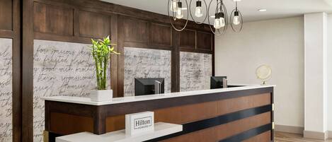 Un mostrador de recepción de hotel con diseño moderno, que presenta un panel de madera con texto escrito a mano, una barra blanca y un cartel que dice "Hilton Honors".