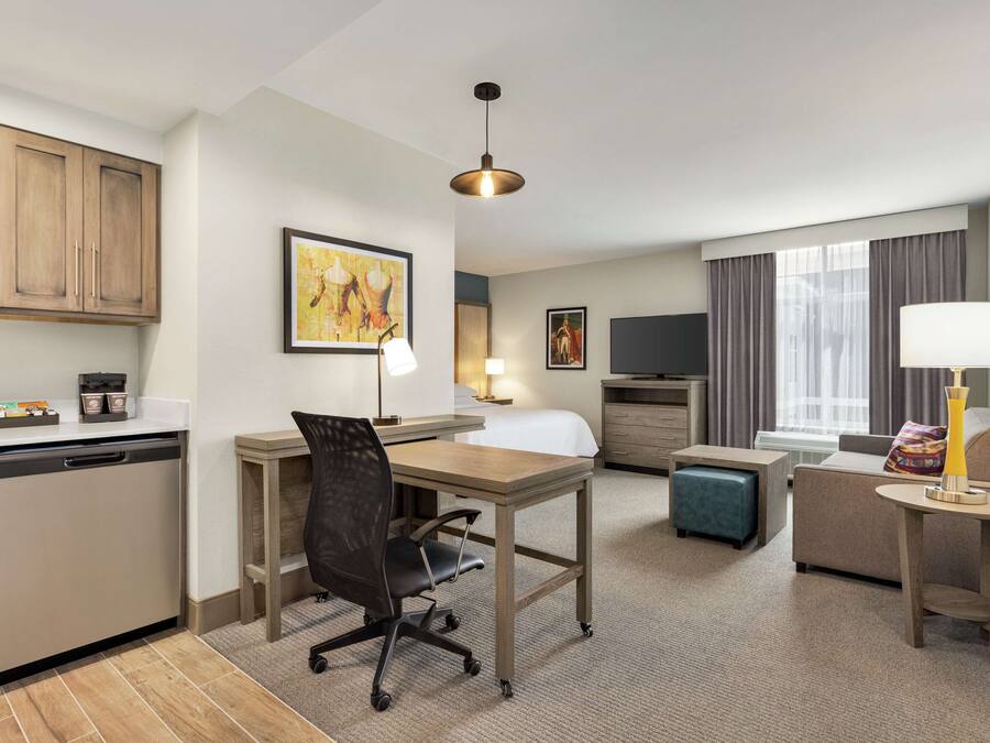 A hotel room with a kitchenette, a dining area, a bed, a TV, and a window with curtains.