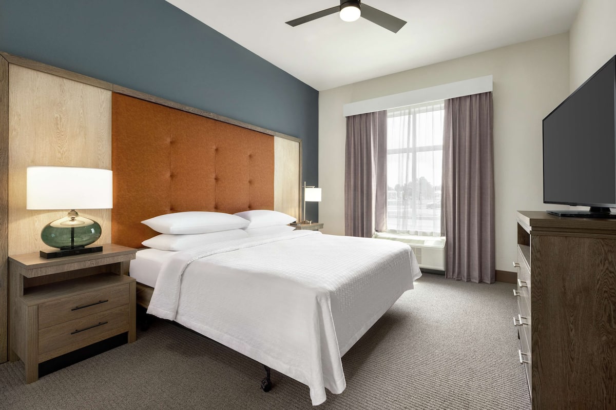 Ein Hotelzimmer mit einem Bett, einem Nachttisch mit Lampe, einem Fernseher und einem Fenster mit Vorhängen.