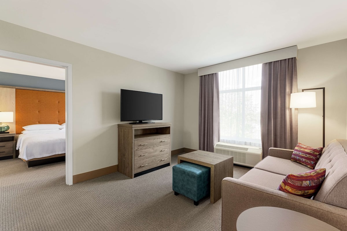 Ein Hotelzimmer mit einem Bett, einem Fernseher auf einer Holzkommode, einer Couch mit Kissen, einem kleinen Tisch und einem Fenster mit Vorhängen.
