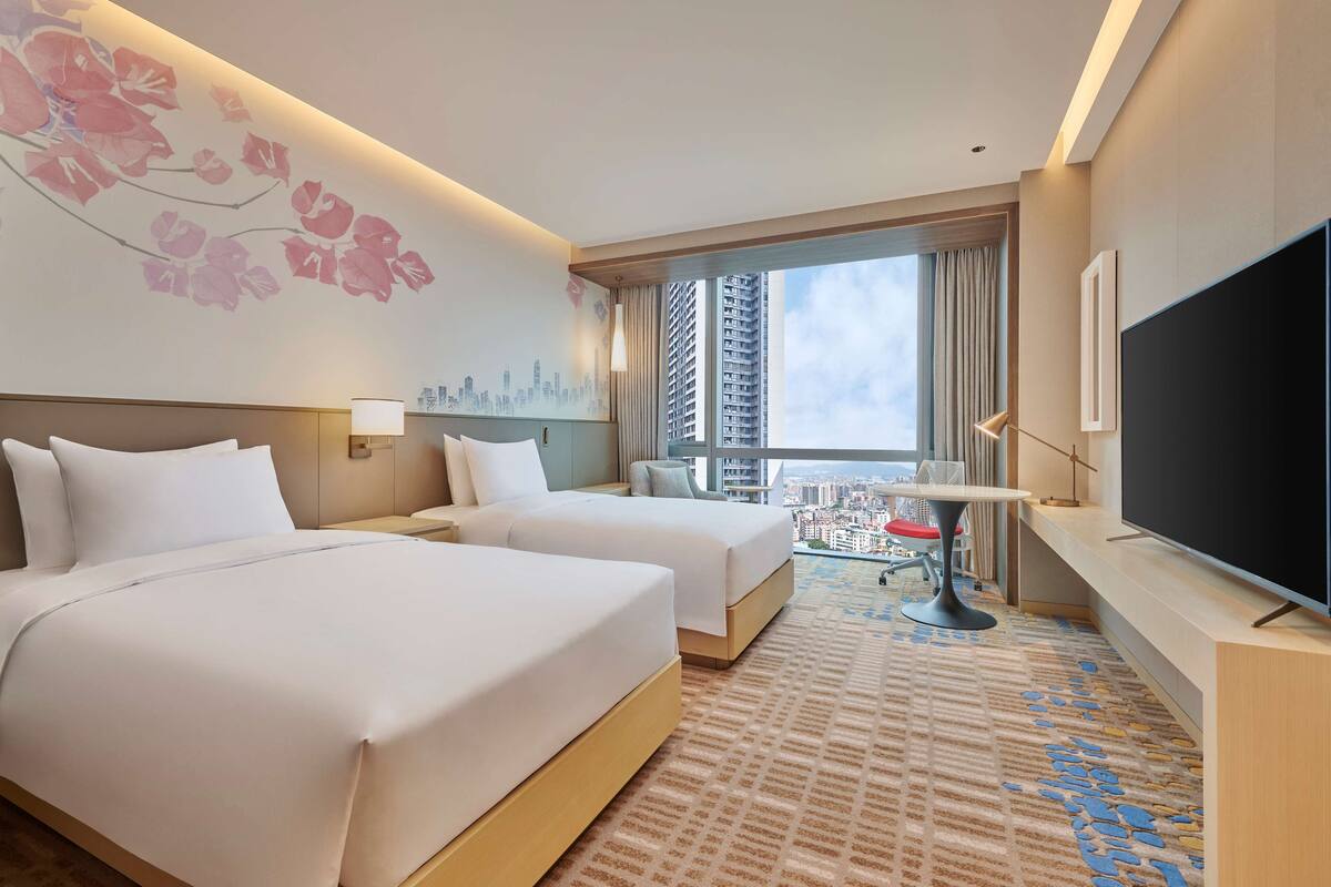 A hotel room with two beds, a TV, a cityscape mural, and a balcony with a city view.