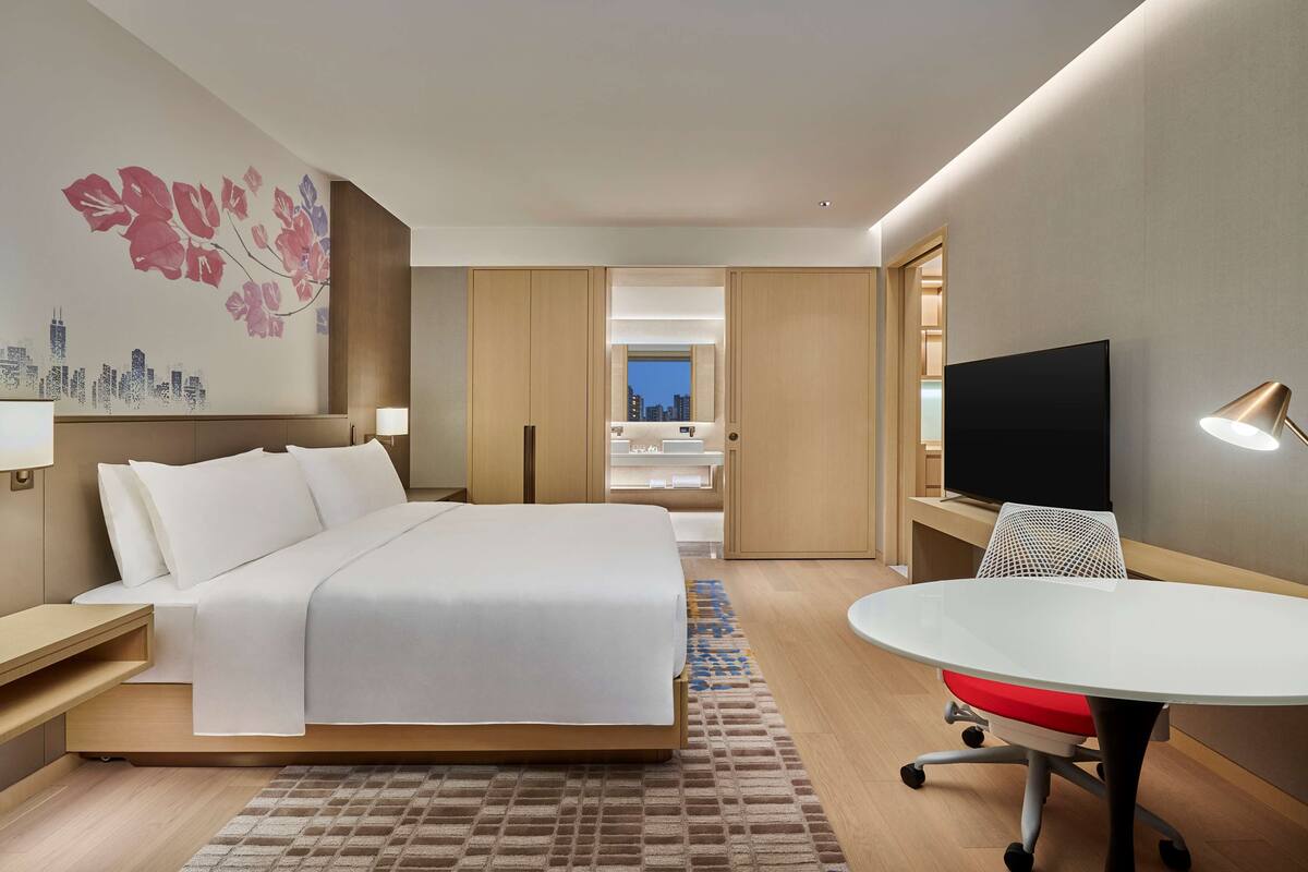 A modern hotel room with a large bed, a desk with a TV, and a view of the cityscape through a window.