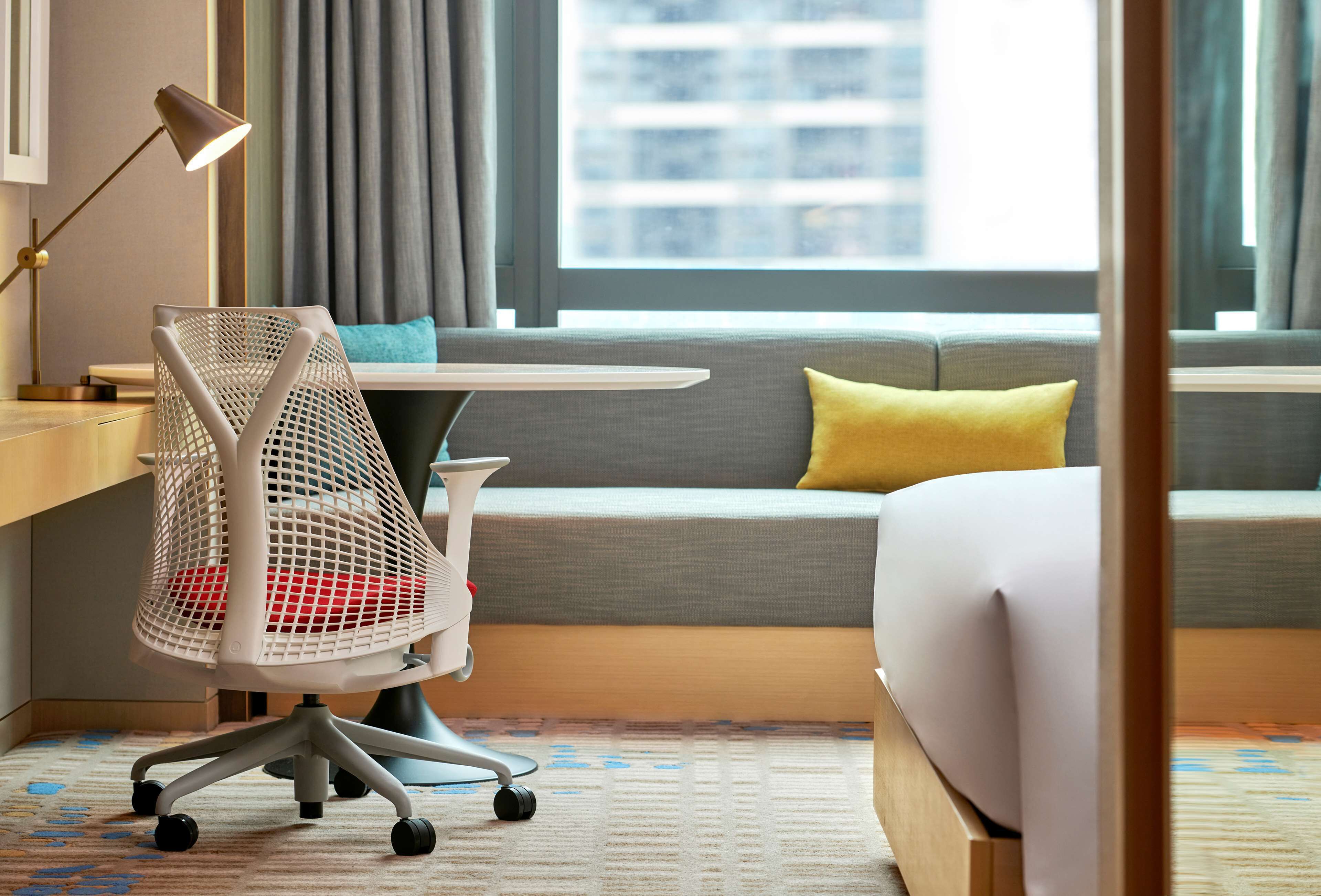 Een hotelkamer met een bed, een bureau met lamp, een stoel, een bank met kussens en een raam met gordijnen.