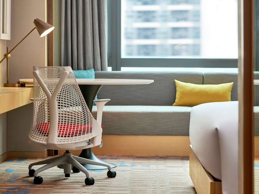 Een hotelkamer met een bed, een bureau met lamp, een stoel, een bank met kussens en een raam met gordijnen.