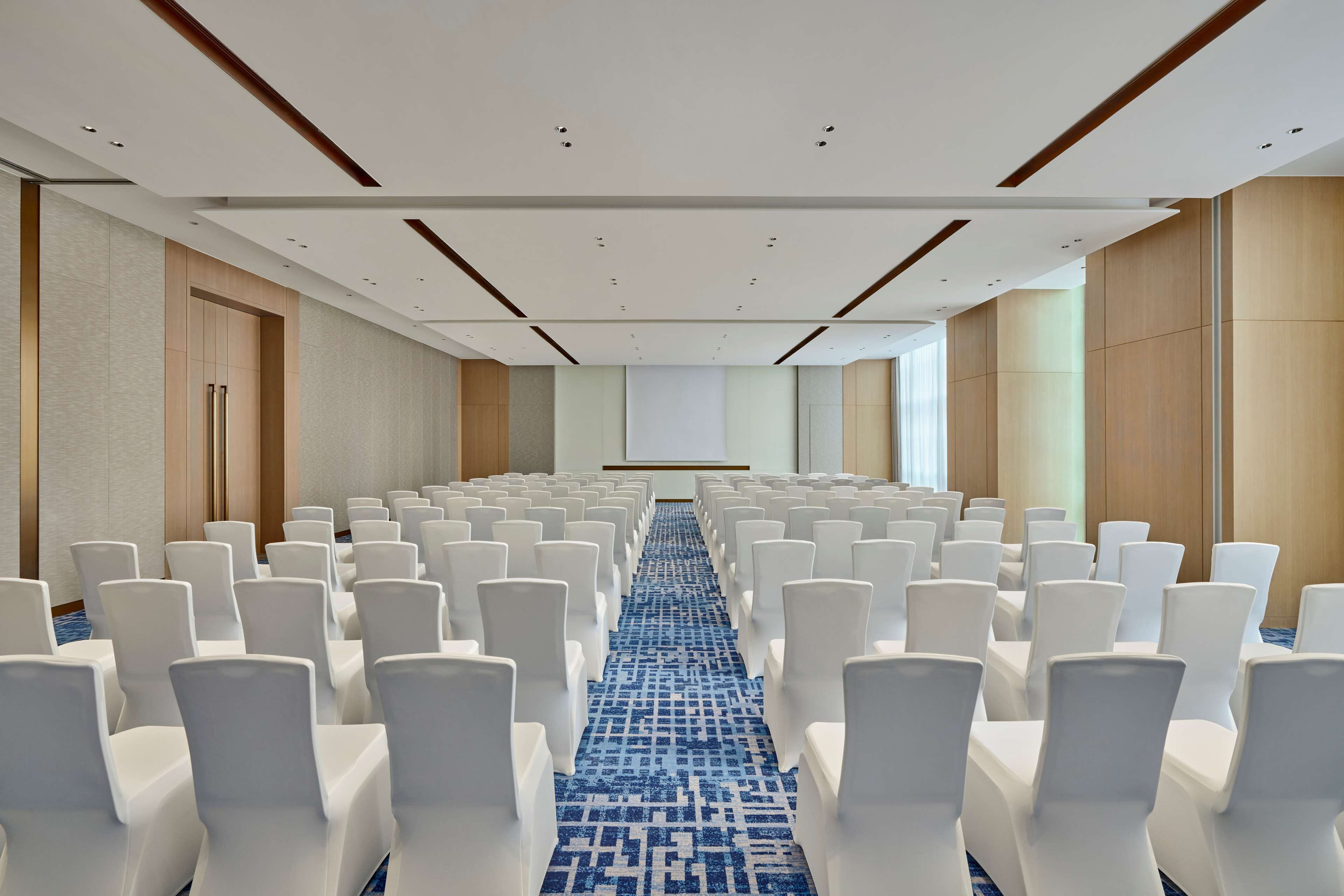 A conference room with rows of white chairs, a blue patterned carpet, and a projector screen at the front.