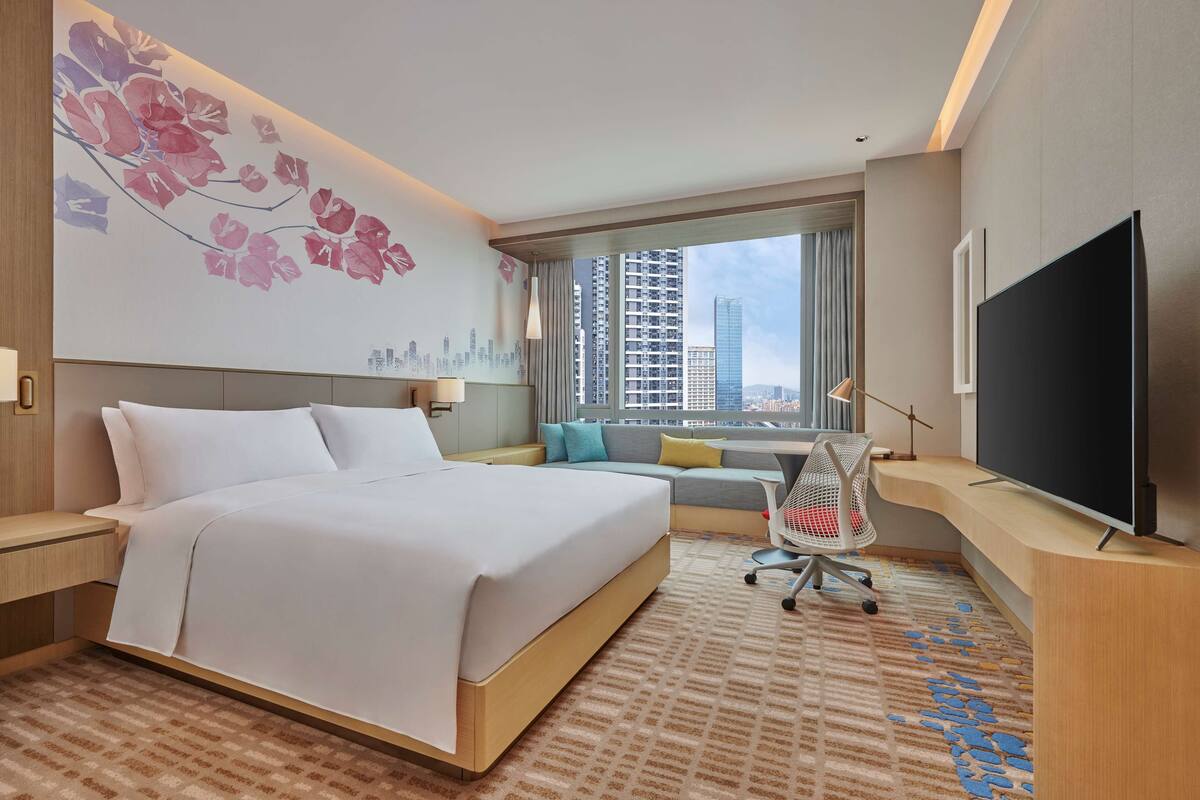 A hotel room with a large bed, a desk with a flat-screen TV, a cityscape mural, and a window with a view of tall buildings.