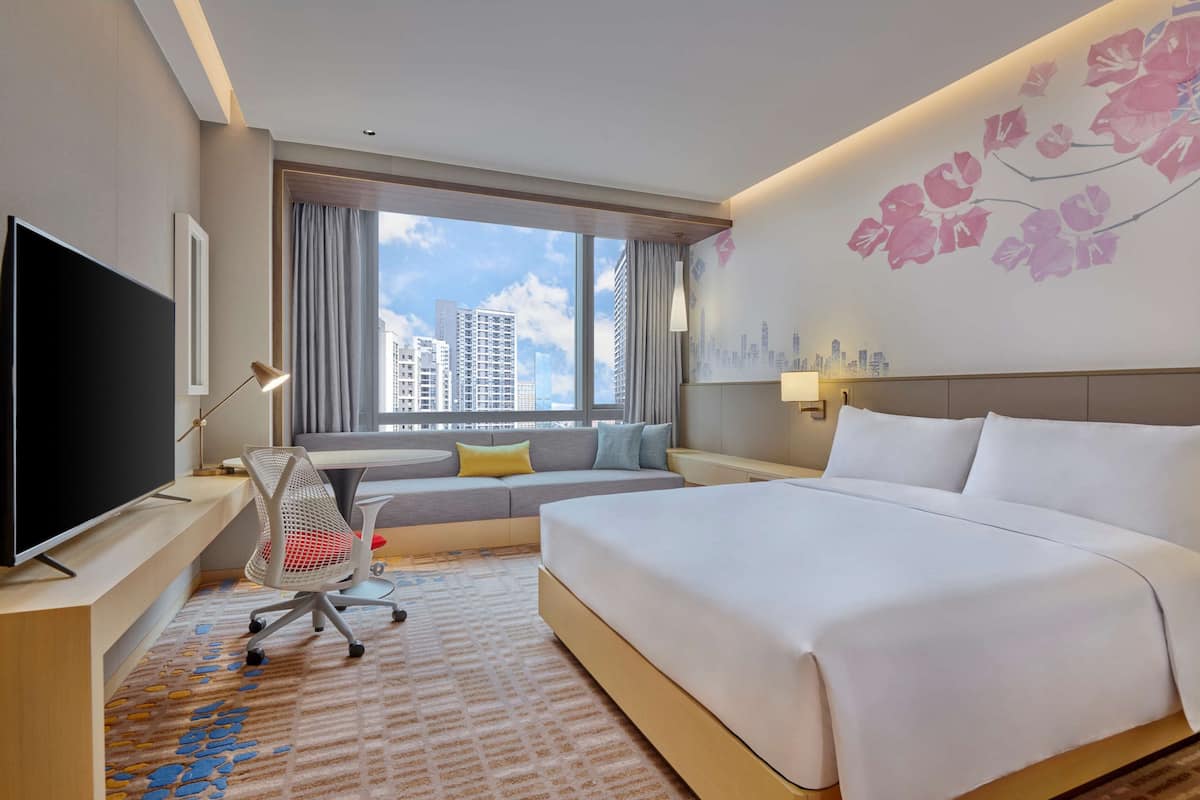 A hotel room with a large bed, a TV, a sofa, and a window with a city view.