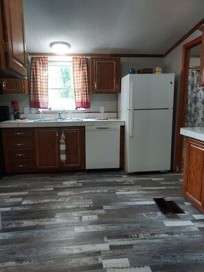 A kitchen with wooden cabinets, a white refrigerator, a dishwasher, and a window with curtains.