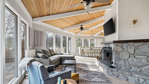 A spacious living room with a stone fireplace, large windows, and a wooden ceiling. - Stylish Modern 6 Bdrm + Hot Tub + Pool Access (Gatlinburg)