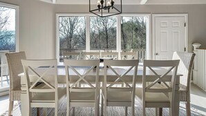 A dining area with a white table and chairs, a geometric ceiling light, and large windows with a view of trees. - Stylish Modern 6 Bdrm + Hot Tub + Pool Access (Gatlinburg)