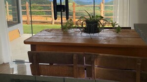 Un coin repas avec une table et des bancs en bois, une plante en pot et une vue sur un paysage montagneux grâce à de grandes fenêtres.