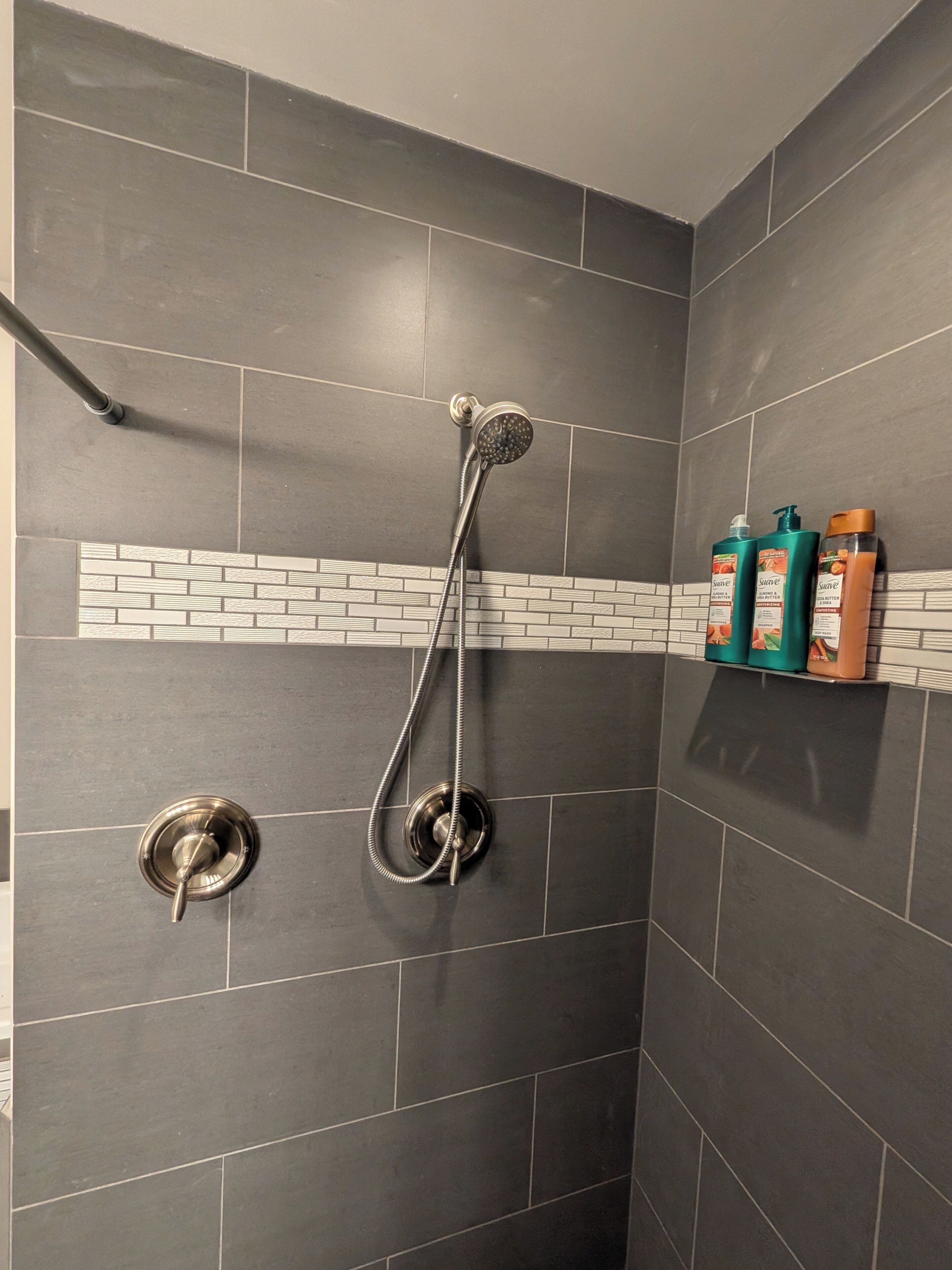 Suihkussa on käsisuihkupää ja seinään kiinnitetty hana. Yksi seinä on laatoitettu valkoisilla tiilikuviolla, ja siinä on hylly, jossa on kolme pulloa hygieniatuotteita.