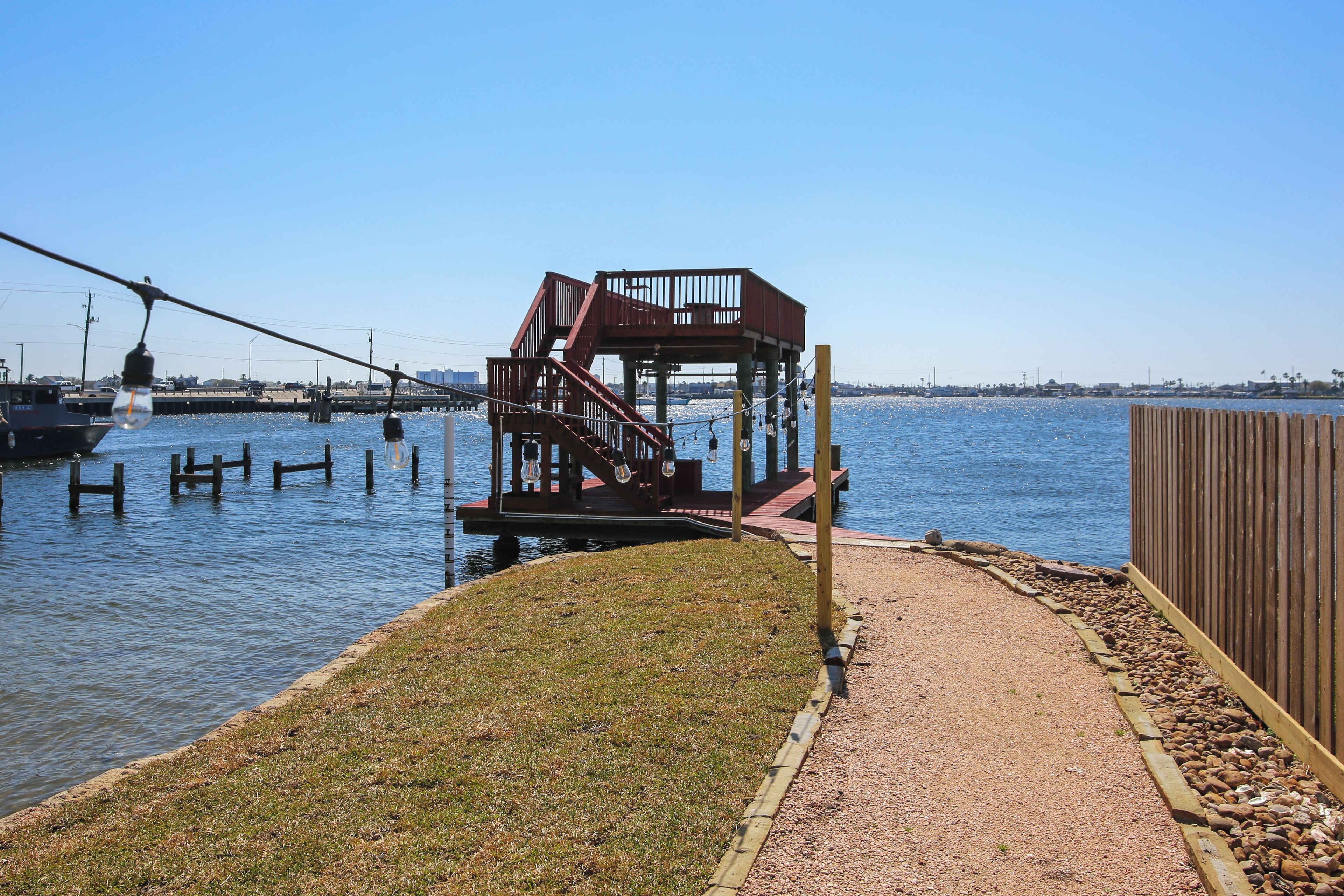 Une terrasse au bord de l’eau, avec un escalier en bois menant à une plateforme, le tout entouré d’un chemin de gravier et d’une clôture en bois.