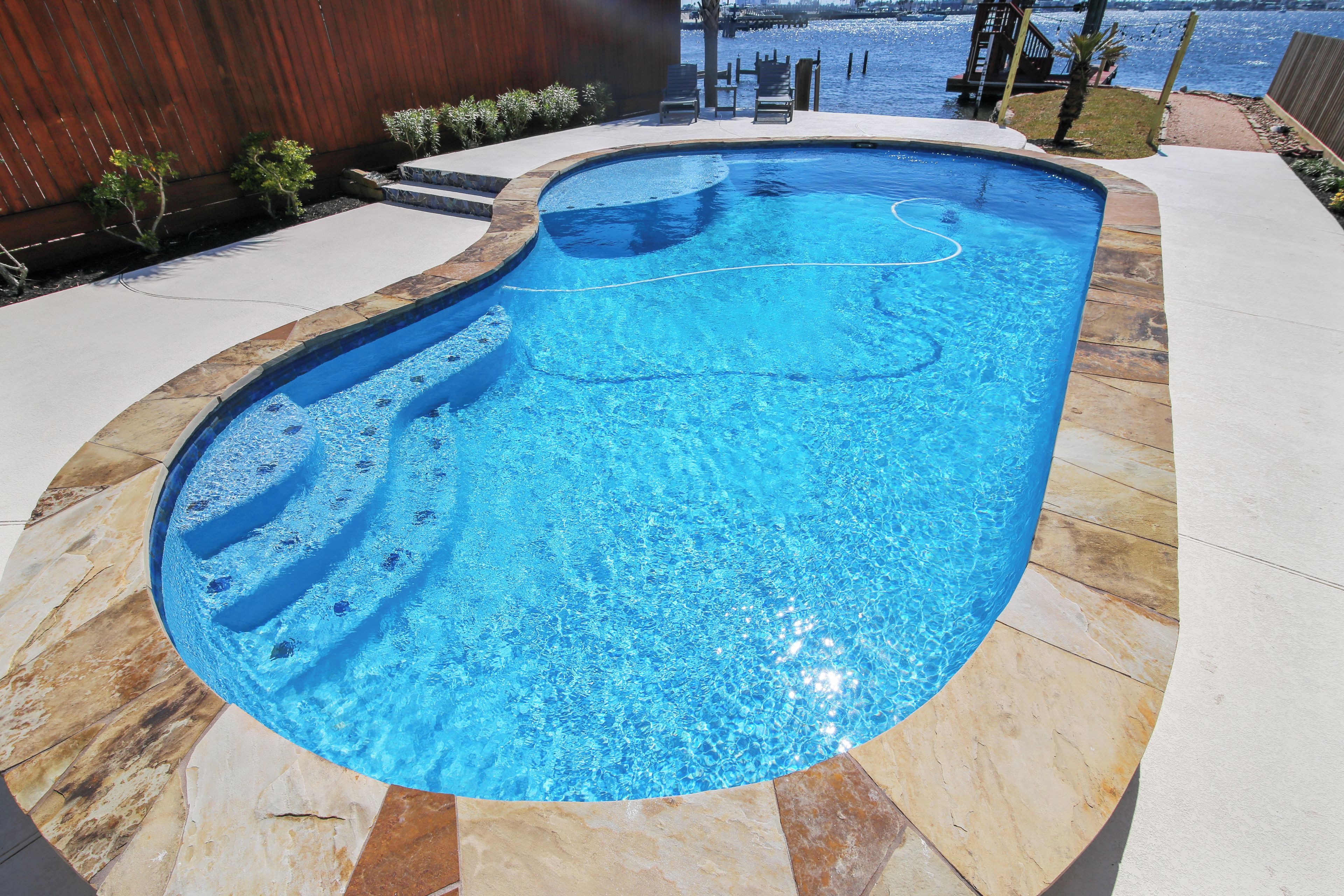 A swimming pool with a curved design, surrounded by a stone deck and a wooden fence.