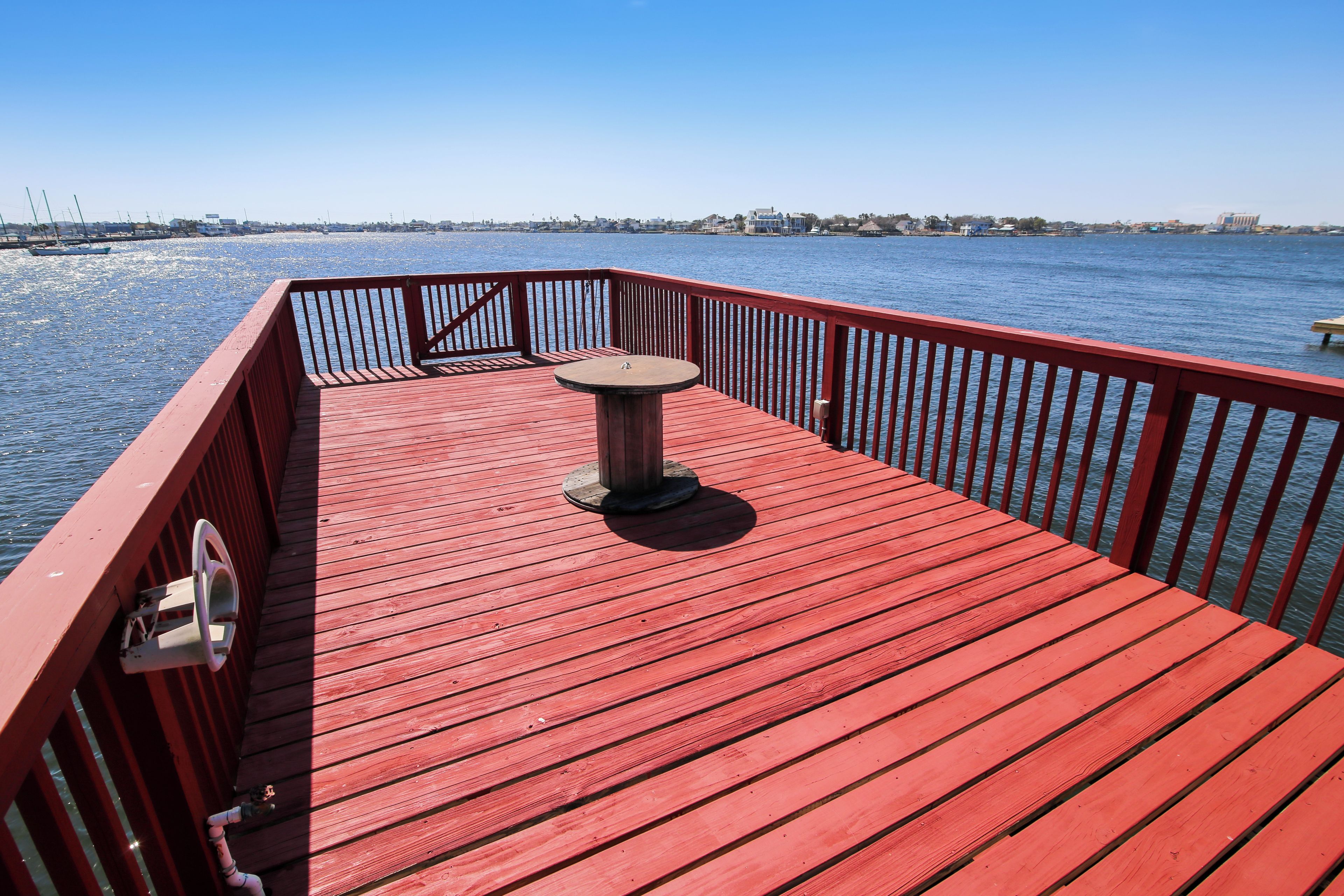 Een rood houten terras met een tafel en een reling, met uitzicht op een waterpartij met boten en gebouwen in de verte.