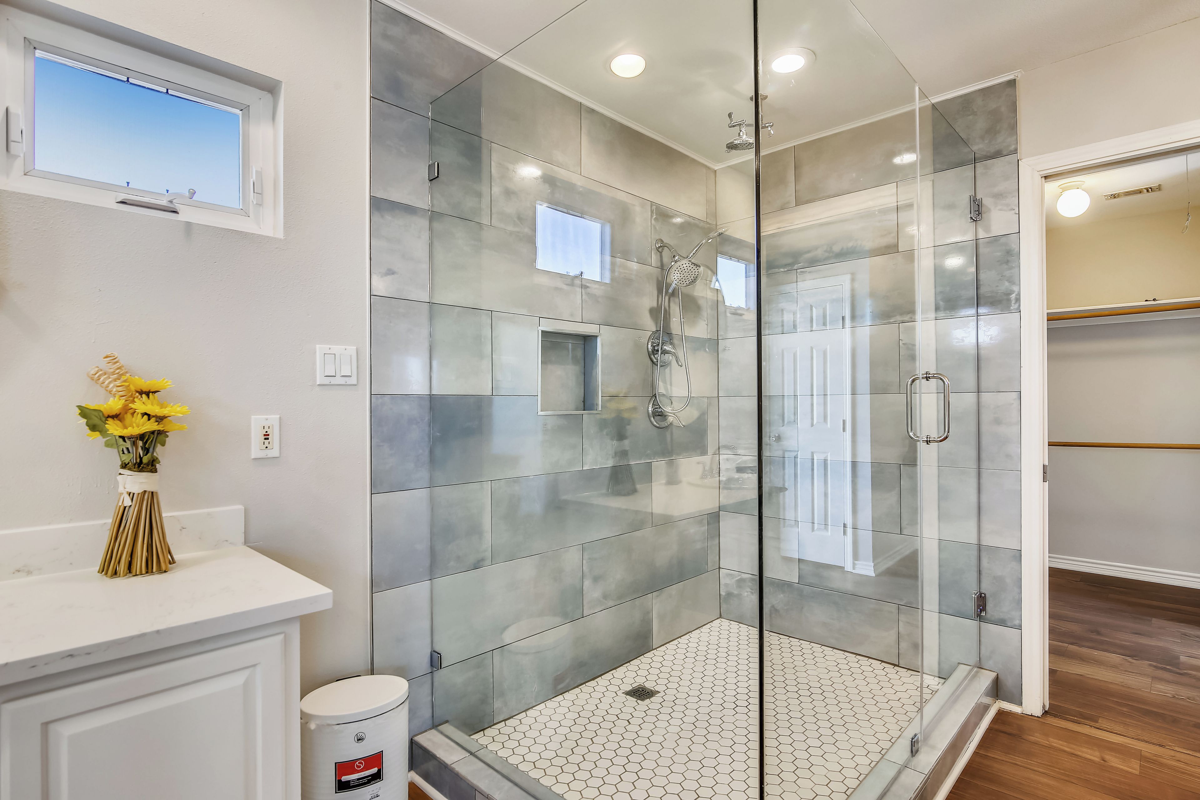 Un baño moderno con ducha separada por vidrio, tocador blanco y una ventana pequeña.