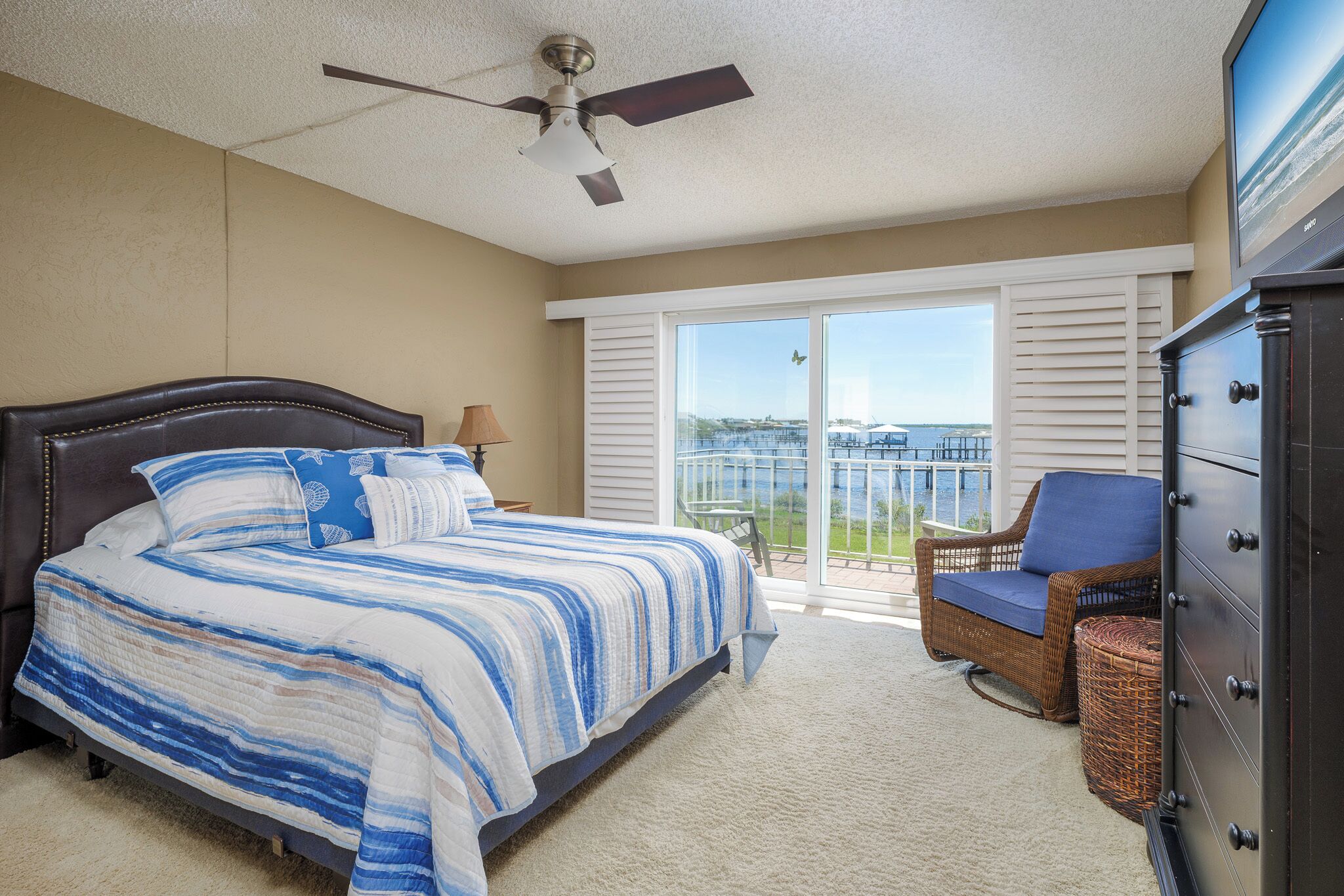 A bedroom with a bed, a ceiling fan, a dresser, a chair, and a view of the outdoors through a sliding glass door.