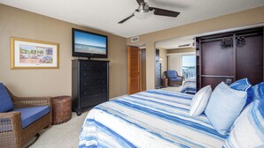 A bedroom with a large bed, a television, a dresser, a wicker chair, and a view of the outdoors. - Point Matanzas B3 by Distinctive Beach Rentals (St. Augustine)