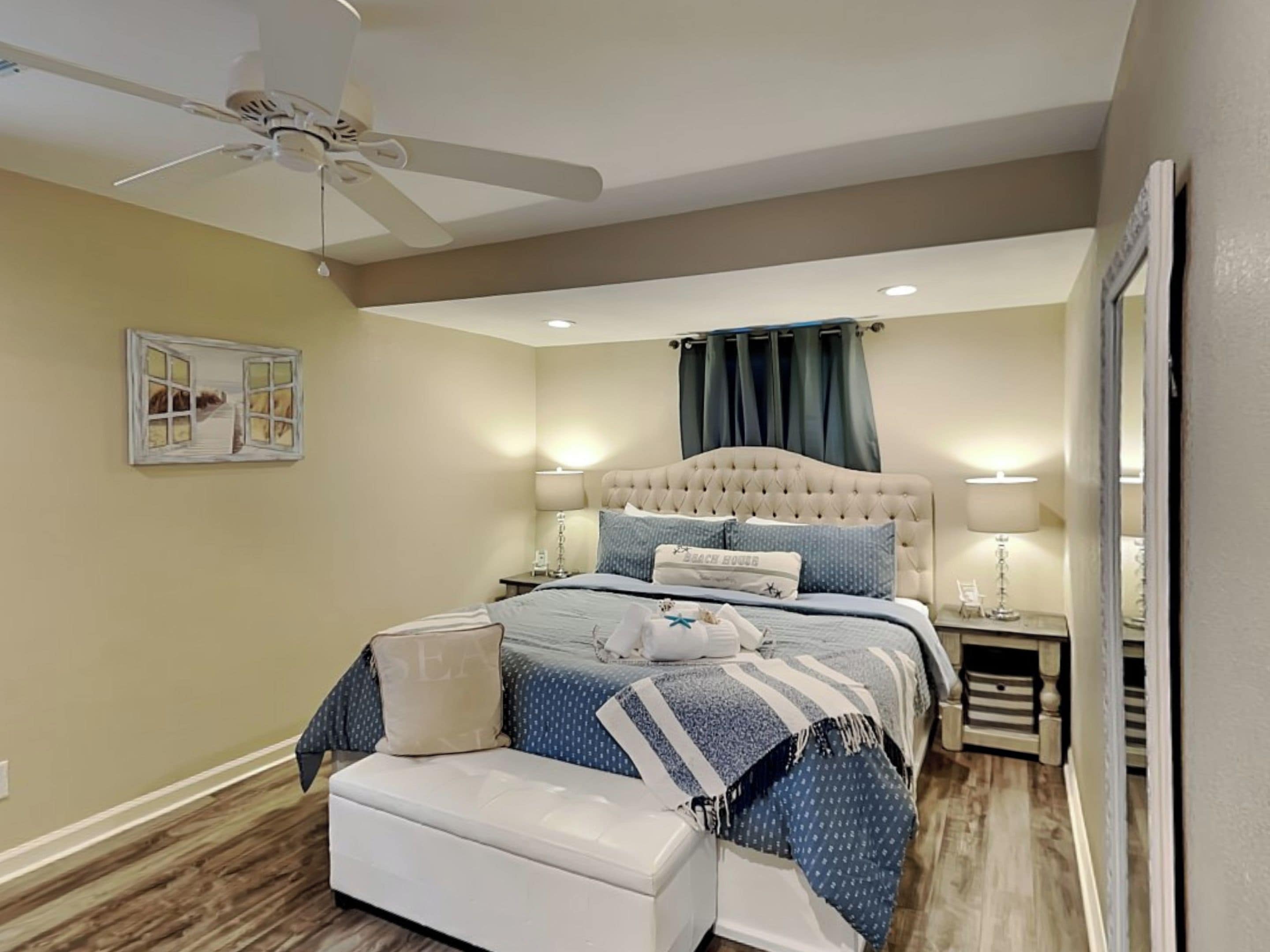 A bedroom with a tufted headboard, a bed with a striped bedspread, a bedside table with a lamp, and a ceiling fan.