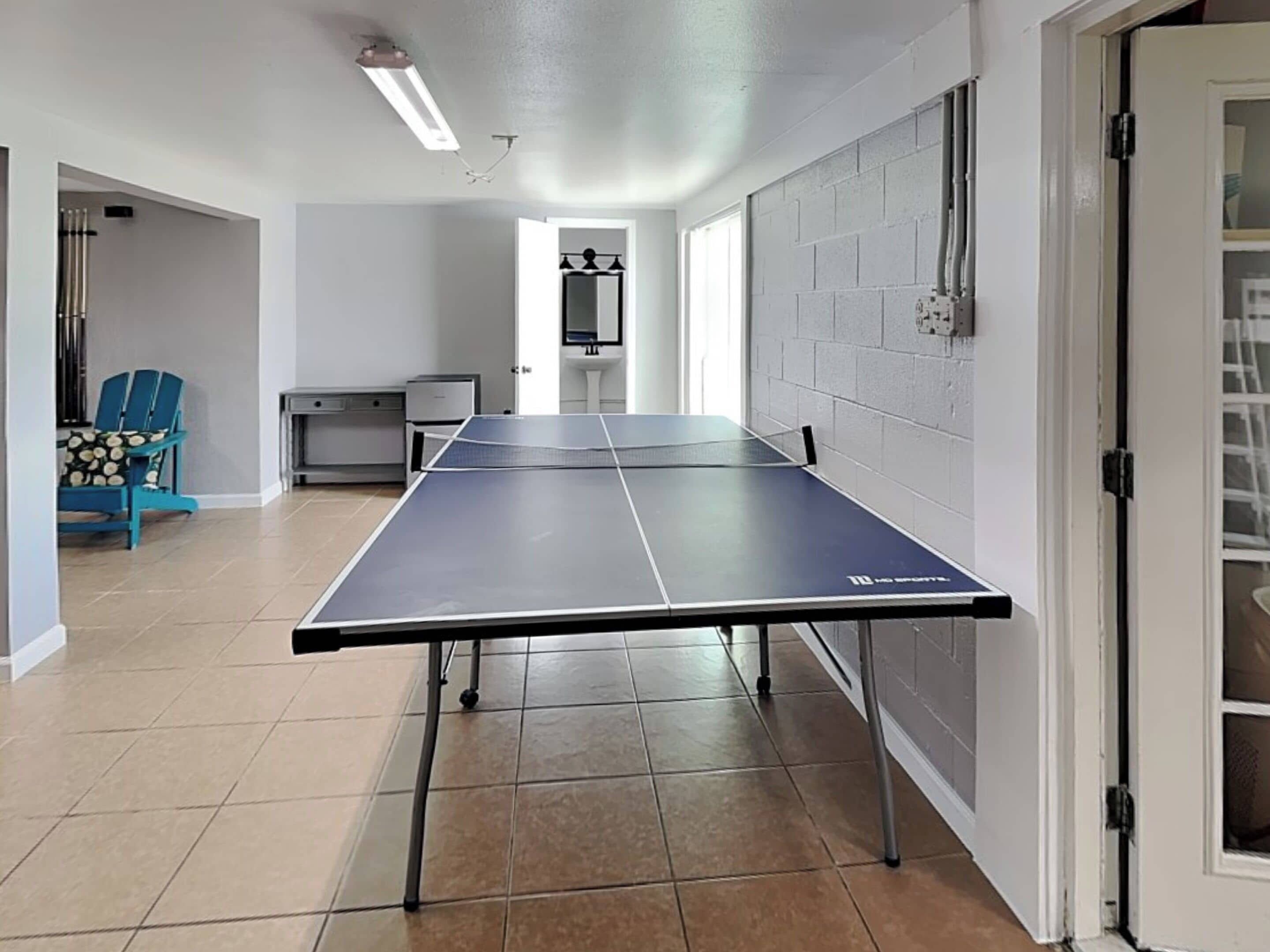 A ping pong table in a room with a tiled floor, a blue chair, a desk, and a white door.