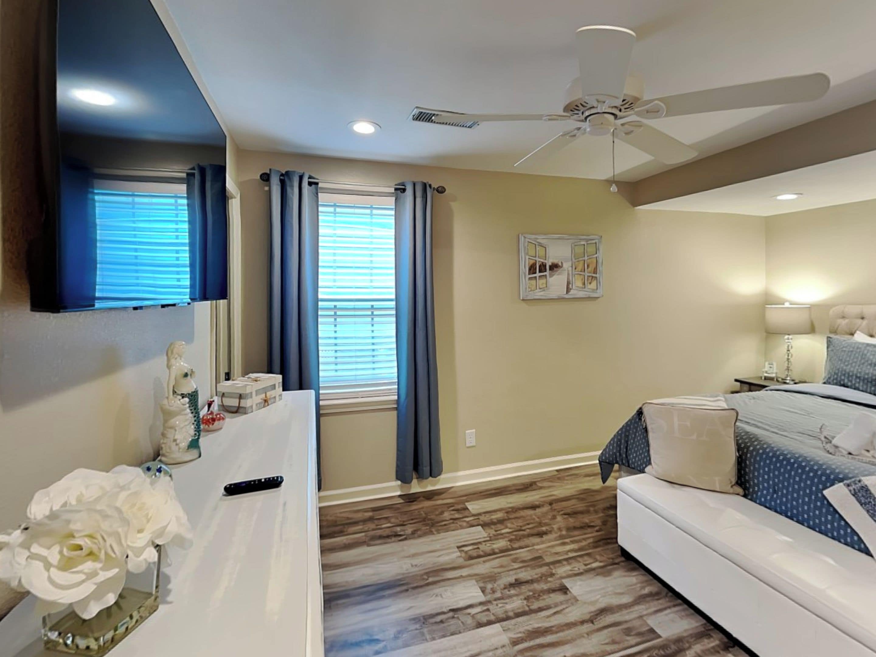 A bedroom with a bed, a ceiling fan, a window with curtains, a wall-mounted TV, and a decorative item on the dresser.