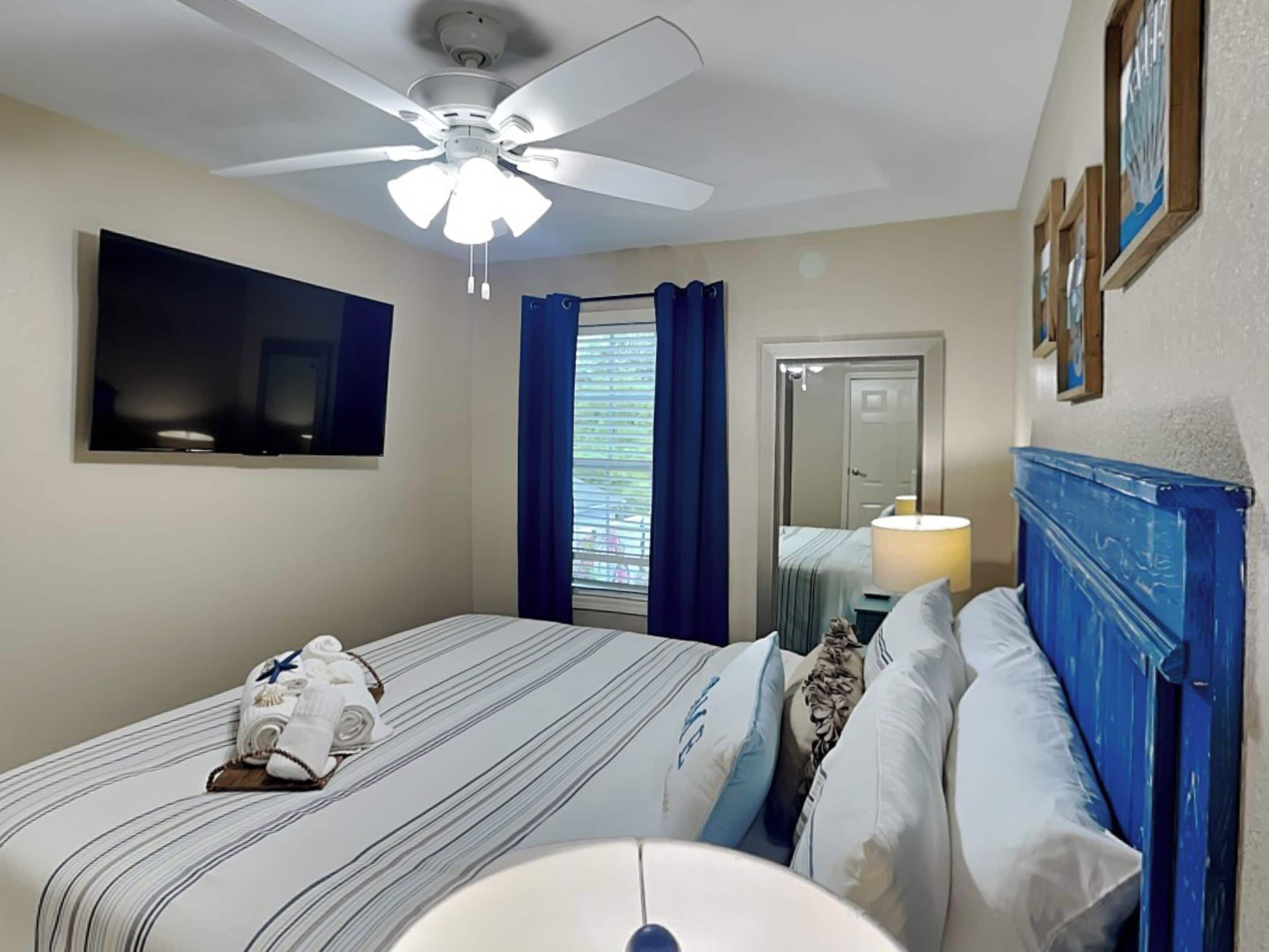 A hotel room with a bed, a ceiling fan, a TV, a window with curtains, and a door.