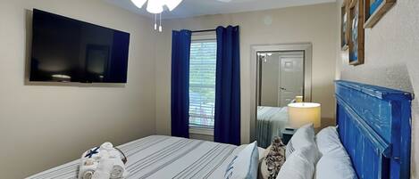 A hotel room with a bed, a ceiling fan, a TV, a window with curtains, and a door.