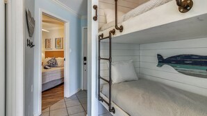 A bunk bed with a decorative fish headboard, a hallway leading to a bedroom with a bed and a door, and a wall with two framed artworks. - Relax by the Beach by Ryson Vacation Rentals (Galveston)