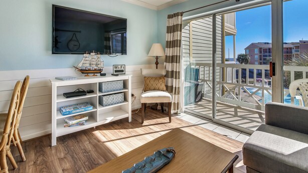 A room with a wooden coffee table, a white armchair, a bookshelf with board games, a small white stool, and a large window with a view of buildings and a pool. - Relax by the Beach by Ryson Vacation Rentals (Galveston)