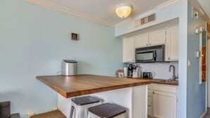 A compact kitchen with white cabinets, a wooden countertop, and a built-in microwave. There are two stools and a small trash can. - Relax by the Beach by Ryson Vacation Rentals (Galveston)