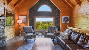 Interior acogedor de una cabaña de troncos con chimenea de piedra, dos sillones con diseños, un sofá de cuero y un amplio ventanal con vistas panorámicas.
