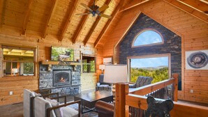 Interior acogedor de una cabaña con chimenea de piedra, vigas de madera, una estatua de oso y un ventanal con vistas panorámicas.