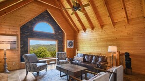 Interior de una cabaña de troncos con un ventanal de arco de piedra, vigas de madera en el techo, chimenea y un amplio ventanal con vistas al exterior.