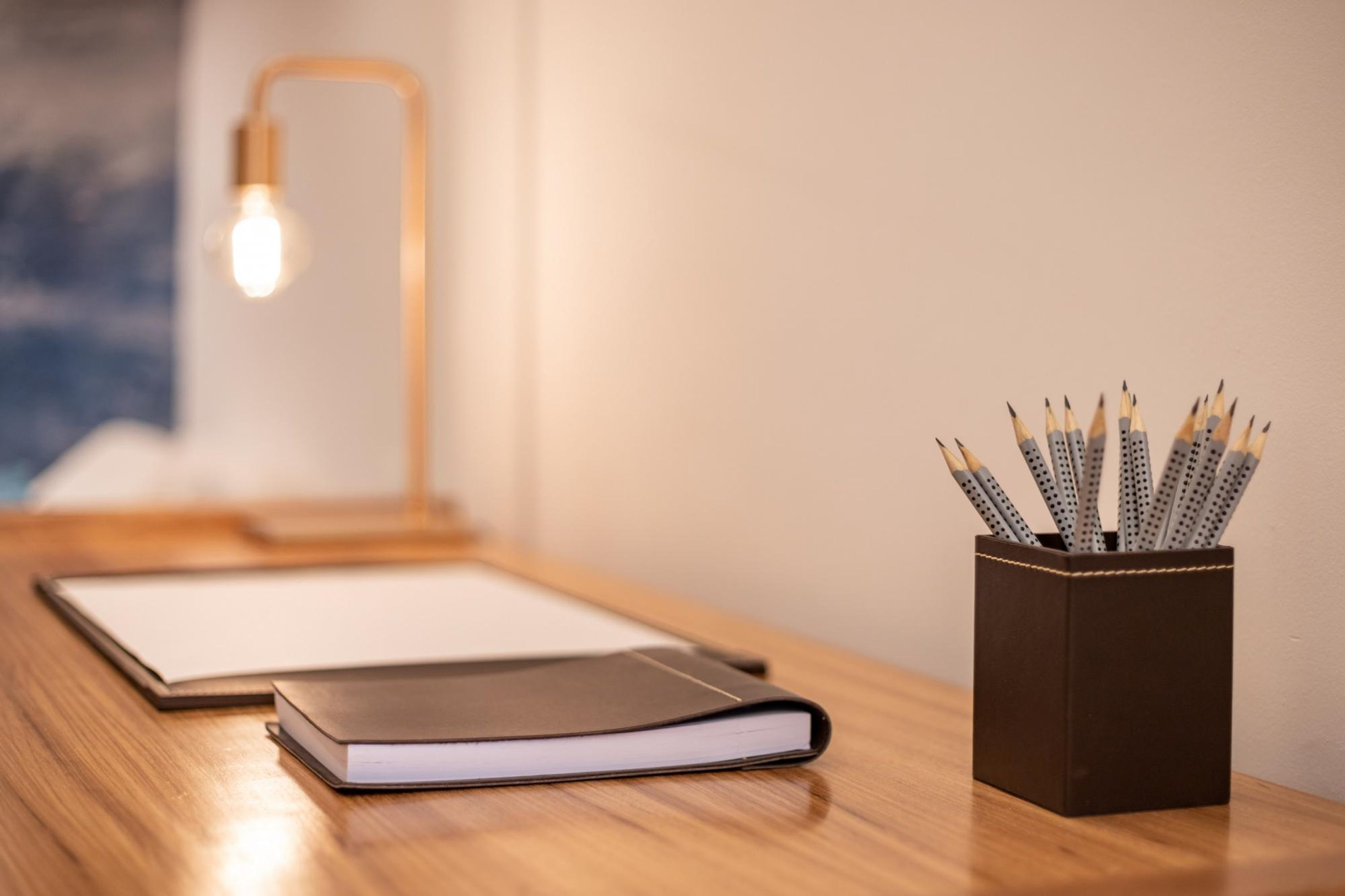 A wooden desk with a notebook, a pen holder with pencils, and a lamp in the background.